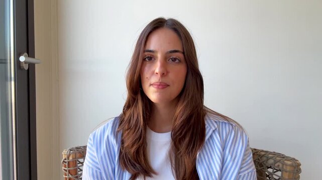 Confident young woman talking directly to the camera during a video conference call, participating in an online interview or webinar from her home office with natural window light