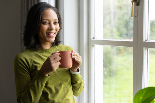 African American woman standing at home by window holding coral mug wearing watch, copy space