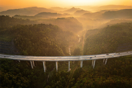 Aerial view of Jalan Tol Bawen - Salatiga bridge spanning over a lush valley with dense forest and misty hills during golden hour, Tengaran, Central Java, Indonesia.