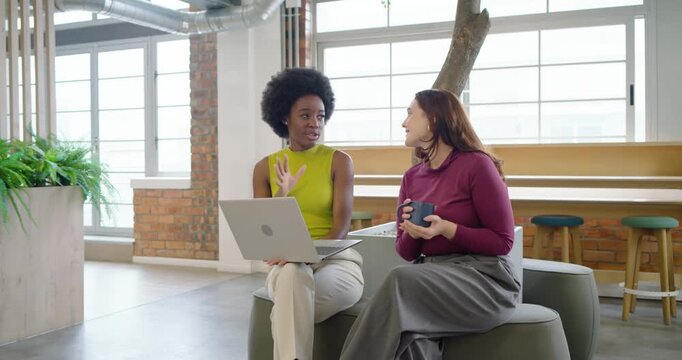 Diverse female coworkers leaning and discussing work on laptop in office with mug after focus pull