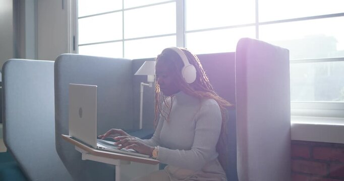 African woman settling into work pod, wearing white headphones, typing on laptop, working remotely