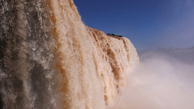 Aerial view of the powerful cascading waters and mist of Iguazu Falls surrounded by lush vegetation under a clear blue sky in Foz do Iguacu, State of Parana, Brazil.