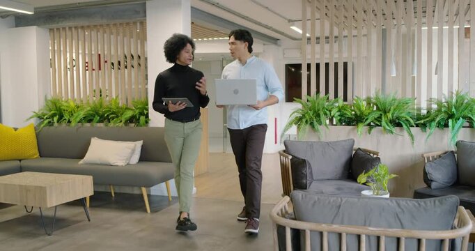 African American and Asian men walking into lounge, discussing project holding tablet and laptop