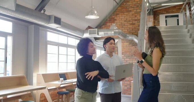 Diverse coworkers watching woman descending stairs presenting tablet to man with laptop in office