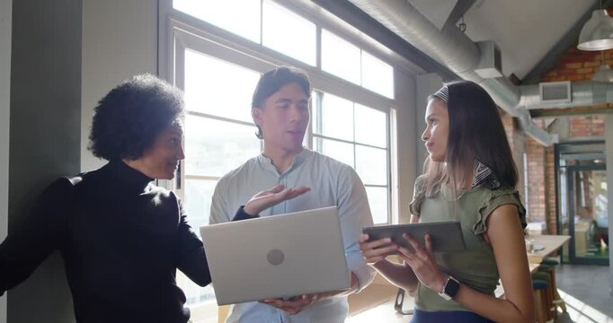 Diverse team being prompted by left one gesturing, pointing at laptop and reviewing tablet in loft