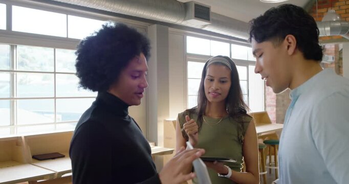 Diverse team approaching tablet holder by windows, exchanging tablet and laptop while reviewing
