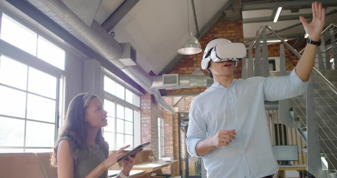 Diverse coworkers reacting to tablet prompts, woman guiding man using VR headset at windows