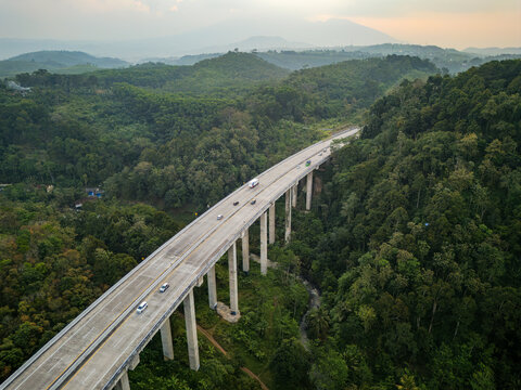 Aerial view of the Jalan Tol Bawen - Salatiga highway bridge spanning lush green hills and forest with vehicles driving along the concrete structure in Sidorejo, Central Java, Indonesia.