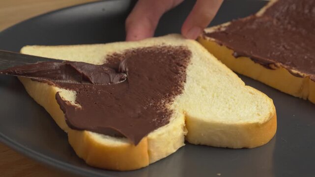 Close up video of hands using a butter knife to spread creamy chocolate hazelnut spread evenly across a slice of white sandwich bread on a black plate