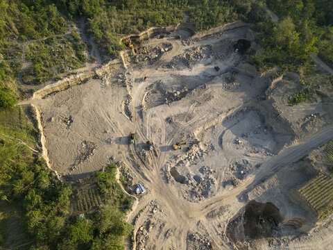Aerial view of Sand and Stone Quarry with heavy machinery and trucks operating in an open-pit mine surrounded by lush vegetation in Manisrenggo, Central Java, Indonesia.