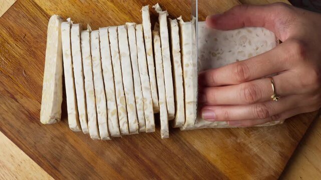 Hands slice firm tempeh on wooden board, top down