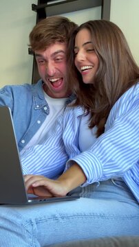 Young smiling couple sitting together on the couch at home while using a laptop computer for online shopping, reacting with surprise and joy to a great offer or good news