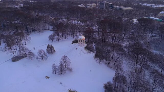Aerial drone shot of Monopteros temple in Englischer Garten, Munich, during a snowy winter sunrise, showing a famous Greek-style landmark and popular tourist destination covered in fresh snow.
