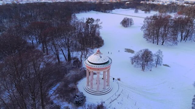 High angle aerial perspective of the historic circular pavilion in Bavaria during winter season, highlighting a popular sledding hill for kids and a scenic viewing point for city tourists.