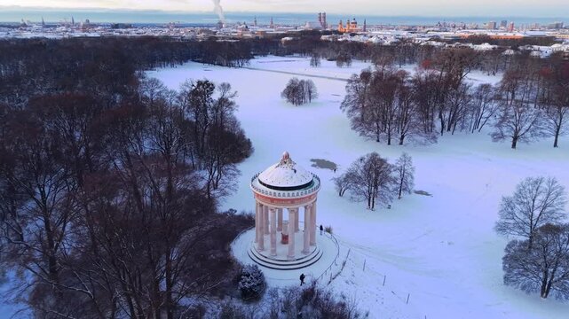 Panoramic winter drone video of the iconic Monopteros temple in Munich, surrounded by white fields and frosty forest, capturing the serene atmosphere of a crisp European sunrise from above.