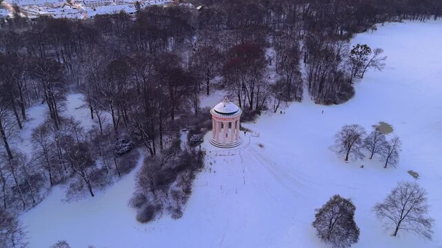 A stunning drone overview of the Monopteros rotunda at dawn, showcasing the Greek-style architecture on a snowy artificial hill within a frozen Englischer Garten park landscape in Munich, Germany.