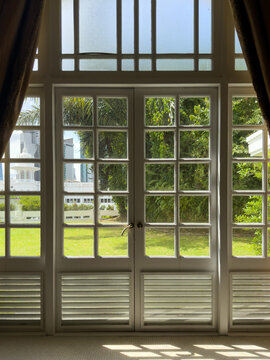 Interior view looking out through classic white wooden French doors with glass panes onto a sunny green garden courtyard