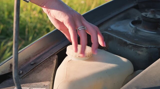 A person opens the fluid reservoir in a vehicle's engine compartment to check levels. This action takes place in an outdoor setting under natural light