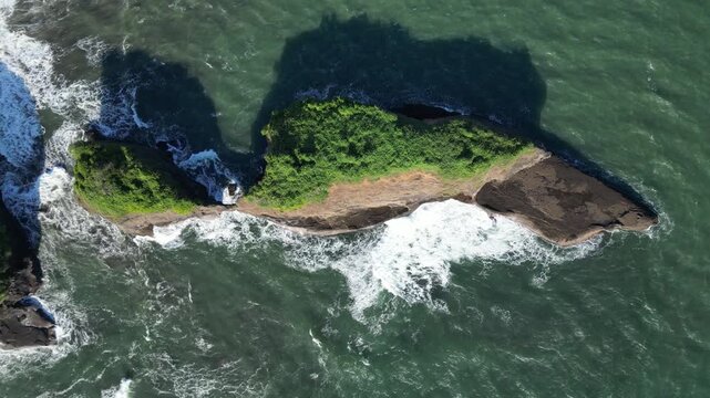 Top down aerial view of a rocky green islet surrounded by crashing ocean waves