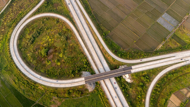 Aerial view of a highway interchange with a loop ramp surrounded by lush green agricultural fields and rice paddies in Indonesia.