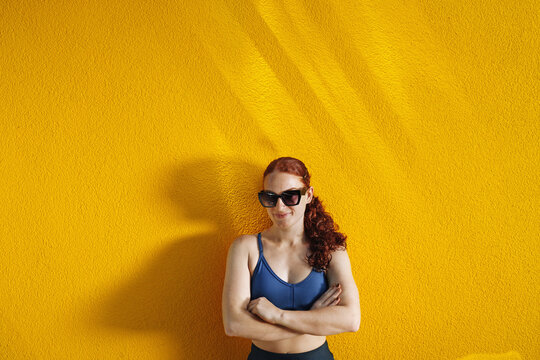 young woman with sunglasses and a sporty look poses in front of a yellow wall. An authentic summer portrait in warm sunlight that embodies sport, a healthy body, and a healthy lifestyle.