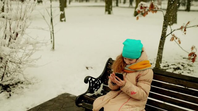 girl sits on bench in winter park with phone
