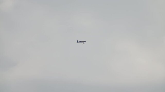 Aerobatic plane in steady level flight, single engine sport aircraft cruising against a hazy grey sky, distant propeller plane at an air show, captured in slow motion