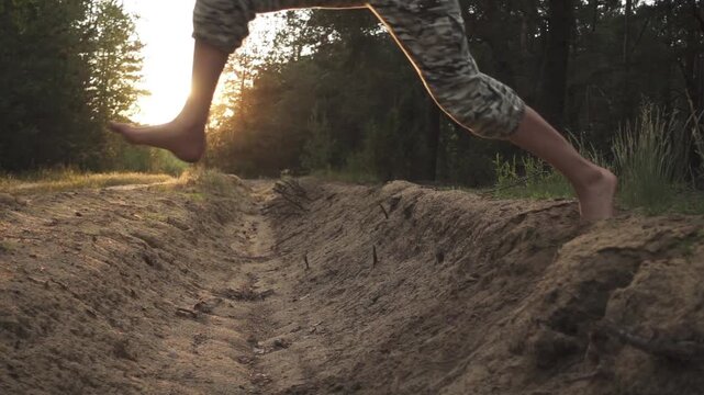 Little girl in camouflage pants jumps through sandy moat in pine forest. Slow motion. Evening time. Sunset.