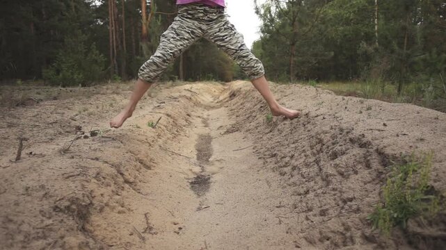 Little girl in camouflage pants jumps through sandy moat in pine forest. Slow motion. Evening time. Sunset.