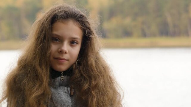 Little girl with long curly loose hair sits on shores of large lake in autumn.
