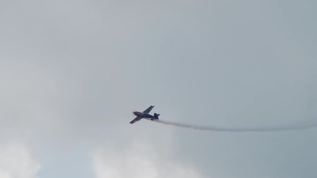 Stunt plane diving toward the left, aerobatic aircraft descending with a thin smoke trail against a grey sky, single engine sport plane at an air show, filmed in slow motion