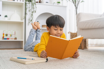 Naklejka premium Asian boy lying on floor reading blank book learning at home, Home education, homeschool, childhood development, self study, elementary student knowledge, smart kid, literature and academic growth