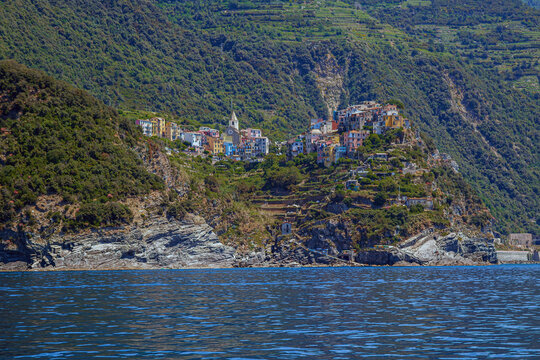 View from the sea of ​​Corniglia, one of the  Cinque Terre villages, a steep cascade of pastel houses descending towards a small harbor carved between dark cliffs. Liguria, La Spezia, Italy.