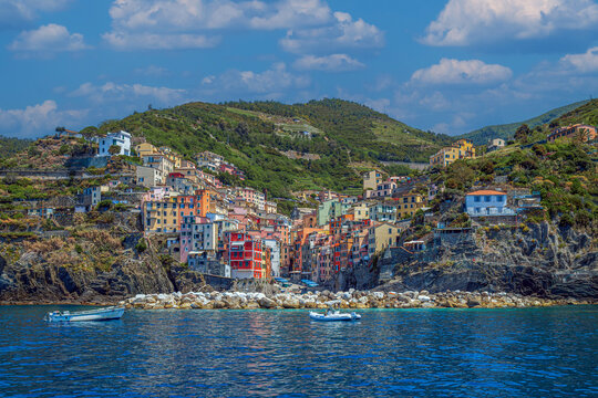 View from the sea of ​​Riomaggiore, one of the  Cinque Terre villages, a steep cascade of pastel houses descending towards a small harbor carved between dark cliffs. Liguria, La Spezia, Italy.
