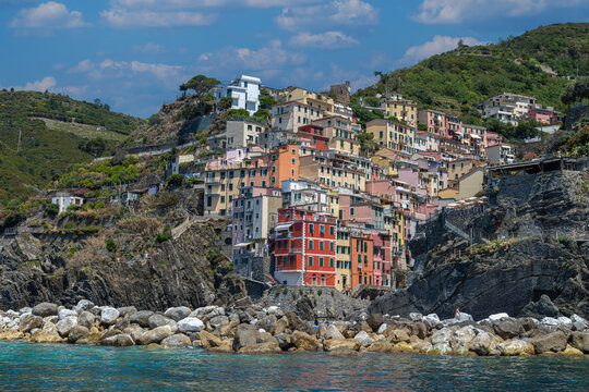 View from the sea of ​​Riomaggiore, one of the  Cinque Terre villages, a steep cascade of pastel houses descending towards a small harbor carved between dark cliffs. Liguria, La Spezia, Italy.
