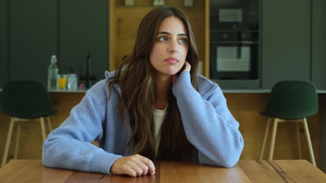 Frustrated and pensive young woman sitting alone at a kitchen table, experiencing boredom and loneliness while lost in thought, showing signs of sadness and contemplation in a modern home