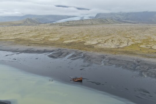 Aerial view of a rusted shipwreck resting on a dark volcanic sand beach with a glacier and mountains in the background near Hofn, Sveitarfelagid Hornaf, Iceland.