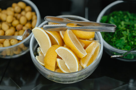 Close view of lemon wedges in glass bowl with tongs, surrounded by potatoes and chopped greens on buffet