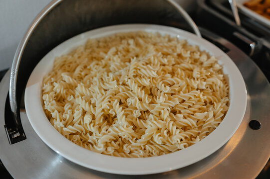 Cooked fusilli pasta fills white serving bowl on buffet cart, showing prepared catering food display bowl