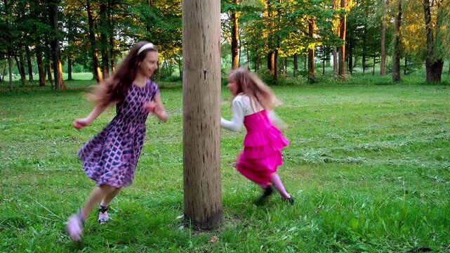 Two beautiful girls sisters running around a wooden post in the summer city park.
