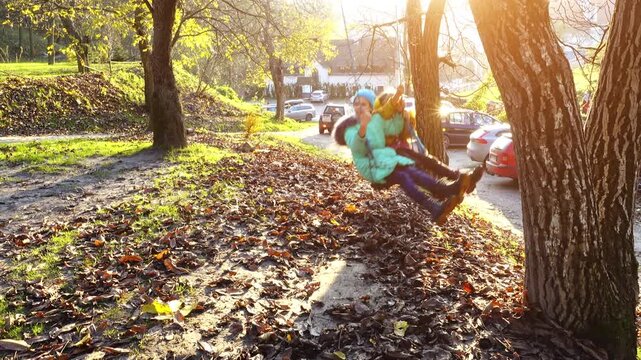 Two little sisters ride a swing, which are suspended with a tree. Autumn nature. Sunny weather.