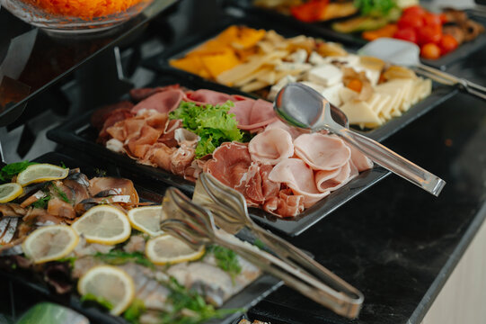 Buffet spread of deli meats, cheeses, smoked fish and lemon garnishes on trays with tongs at catering ham