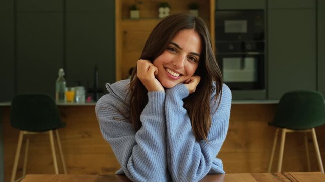 Authentic portrait of a beautiful young woman smiling and having fun at home while sitting at a wooden table in her modern kitchen, showing her genuine happiness and carefree lifestyle