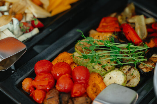 Close view of roasted vegetables cherry tomatoes, zucchini and greens in serving tray beside metal tongs