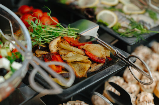 Close up view of grilled vegetables, herbs and tongs arranged on buffet trays for restaurant service now