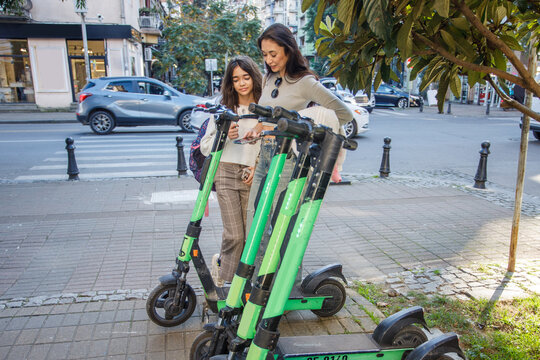 Mother with girl stand beside e-scooters on a city street while looking at their phones in the afternoon