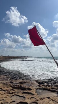 Red warning flag flying on a windy beach above waves, coastal safety, dangerous swimming conditions, seaside travel, ocean awareness, summer vacation.