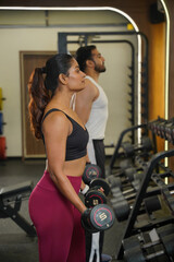 Fototapeta premium Maharashtra, Sion, India- 22nd March 2026- Young Indian girl and guy doing workout with dumbbells, looking into the mirror in gym.