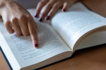 Fototapeta premium Person's hand with painted fingernails pointing finger along lines of text in an open book, symbolizing learning, education, reading, and knowledge