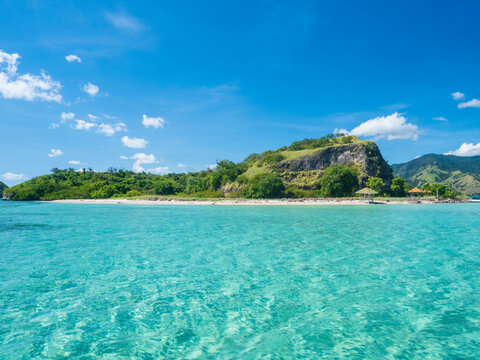 Crystal clear turquoise waters surround a small tropical island in Riung 17 Islands National Park, Indonesia. A peaceful beach, lush green hills, and blue skies create a serene paradise